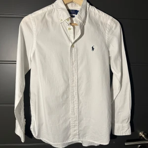 Vit skjorta från Polo Ralph Lauren - Klassisk vit skjorta från Polo Ralph Lauren med blå broderad logga på bröstet. Skjortan har lång ärm, knappar framtill och button-down krage. Perfekt för en stilren och tidlös look. Tröjan är i en slim model och tröjan är i väldigt bra skick, knappt använd. Skjortan är i storlek 160.