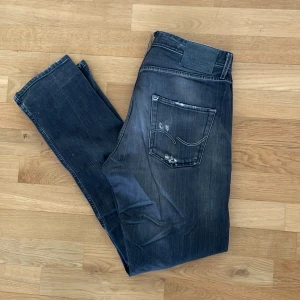 Gråa  jeans med slitningar - Säljer ett par svarta jeans från Jack and Jones modell Erik Anti Fit. Jeansen har en tvättad look med slitningar och små hål på låret samt bakfickorna. Klassisk femficksmodell med knappgylf och raka ben.