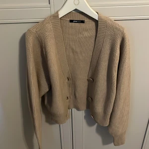 Beige stickad kofta från Gina Tricot - Säljer en beige stickad kofta från Gina Tricot med ribbad struktur. Koftan har långa ärmar, djup v-ringning och stora knappar framtill. Perfekt att slänga över en topp för en chill look.