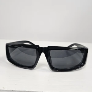 Svarta rektangulära solglasögon - Snygga svarta solglasögon med rektangulära bågar och mörka glas. Glasögonen har en modern, kantig design och breda skalmar som ger en cool och futuristisk look. Perfekta för dig som vill sticka ut med en trendig accessoar.