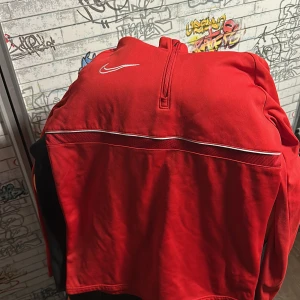 Röd Nike Dri-Fit tröja med halv dragkedja - Säljer en röd Nike Dri-Fit tröja med vit broderad Swoosh-logga på bröstet och halv dragkedja. Tröjan har en vit detaljlinje över bröstet och är perfekt för träning eller sportiga outfits.