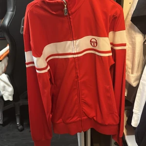 Röd track jacket från Sergio Tacchini - Röd track jacket från Sergio Tacchini med vita ränder och broderad logga på bröstet. Jackan har dragkedja framtill, ståkrage och ribbade muddar. Perfekt för en sportig och retro stil.