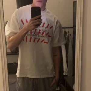 Vit t-shirt från Maison Margiela  - Vit t-shirt från Maison Margiela med röd och svart grafisk text och tryck framtill. Klassisk passform och rund halsringning. Perfekt för dig som gillar stilrena plagg samt grym till sommaren!