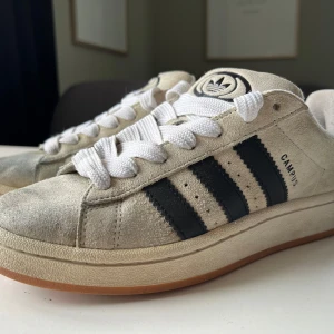 Adidas Campus 00s W sneakers i beige och svart - Säljer ett par Adidas Campus 00s W sneakers i beige mocka med svarta ränder och vit sula. Skorna har klassisk Adidas-logga på plösen och hälen samt breda vita skosnören. Perfekta för dig som gillar retrostil!