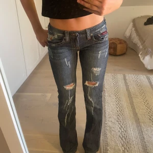 Blå bootcut jeans från Lady Witch - Säljer ett par blå bootcut jeans från Lady Witch med slitningar och broderad orange katt på bakfickan. Jeansen har låg midja och är bootcut. W27, jag är 176 cm lång