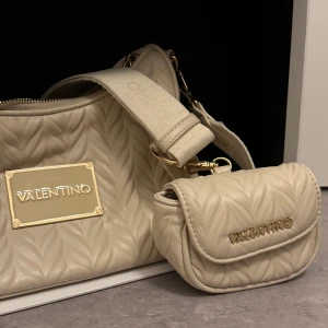 Beige crossbody väska - Säljer en beige axelväska från Valentino med quiltad design och gulddetaljer. Väskan har en bred axelrem och en matchande liten väska som kan fästas på remmen. Perfekt för att bära det viktigaste med stil. Är väldigt öppen med prisförslag och vi kan säkert komma överens! Det är bara att skicka bud!
