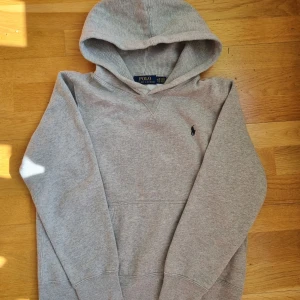 Grå hoodie från Polo Ralph Lauren - Säljer en klassisk grå hoodie från Polo Ralph Lauren med broderad logga på bröstet. Tröjan har huva och långa ärmar, perfekt för en avslappnad stil. Mjuk och bekväm med ribbade muddar vid ärmslut och nederkant.