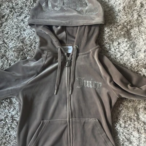 Beige velour hoodie från Juicy Couture - Säljer en jättefin beige velour/brun hoodie från Juicy Couture med glittrande stenar på bröstet och luvan. Jackan har dragkedja framtill, fickor och långa ärmar. Perfekt för en avslappnad och trendig look.