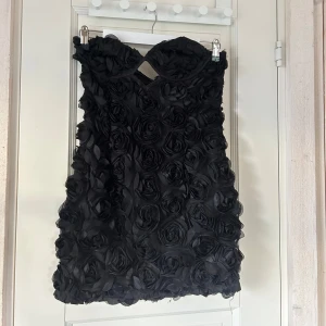 Svart rosmönstrad klänning från PrettyLittleThing - Supersnygg svart klänning från PrettyLittleThing med ros-mönster och utskärning vid bysten. Perfekt för fest och sommaren! Axelbandslös och kort modell. Helt ny!!!!🩷 lappar kvar. 