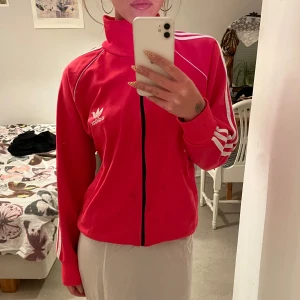 Rosa Adidas track jacket med vita ränder - Säljer en rosa Adidas track jacket med vita ränder på ärmarna och vit logga både fram och bak. Jackan har dragkedja hela vägen, hög krage och lång ärm. Klassisk sportig stil med bekväm passform.