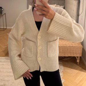 Cremevit stickad kofta från Gina tricot  - Mysig vit stickad kofta från Gina tricot med djup v-ringning och stora knappar framtill. Koftan har två framfickor och breda ribbade muddar vid ärmslut och nederkant.❤️