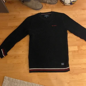 Mörkblå stickad tröja från Bondelid - Säljer en mörkblå stickad tröja från Bondelid med broderad logga på bröstet. Tröjan har långa ärmar och ribbade muddar med röd och vit rand vid ärmslut och nederkant. Perfekt för kyliga dagar!