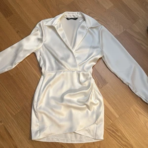 Vit omlottklänning i satin - Säljer en elegant vit omlottklänning i satin med lång ärm och djup v-ringning. Klänningen har en figurnära passform och kort längd, perfekt för speciella tillfällen. Snygg krage och lätt glansigt material ger en lyxig känsla.