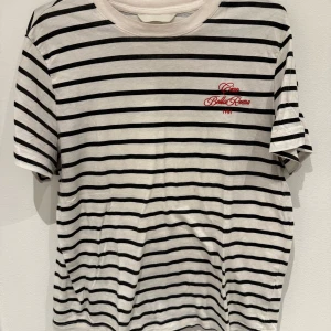 Randig t-shirt  - Säljer en vit och svart randig t-shirt med broderad röd text på bröstet. T-shirten har rund halsringning och korta ärmar. Perfekt för en avslappnad stil.
