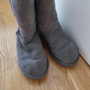 Gråa boots med fårullsfoder - Mysiga grå boots med rund tå och platt sula. Skorna är fodrade med mjuk fårull på insidan och har en klassisk, enkel design. Perfekta för kyliga dagar när du vill hålla fötterna varma.