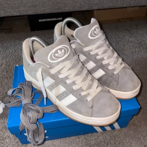 Grå Adidas Campus  - Säljer ett par grå Adidas Campus 00s sneakers i mocka med vita ränder och vit sula. Extra skosnören i grå medföljer. Perfekta för dig som gillar streetwear och vill ha en stilren look. Köptes förra året för ca 1600 kvitto finns💕