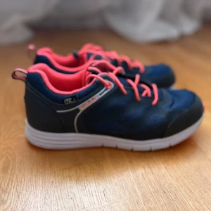 Blå och rosa sneakers med vit sula - Säljer ett par snygga blå sneakers med rosa detaljer och snörning. Skorna har en vit, platt sula och är perfekta för vardagsbruk eller träning. Ovandelen är i mesh-liknande syntetmaterial för bra andningsförmåga.