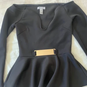 Svart klänning med gulddetalj från NLY One - Snygg svart klänning från NLY One med lång ärm, markerad midja och guldfärgat bälte. Klänningen har en v-ringad urringning och figurnära passform upptill med utsvängd kjol.