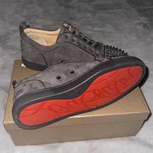 Grå mockasneakers med nitar Louboutin  - Säljer ett par grå sneakers i mocka med svarta nitar på tån och snörning. Skorna har en klassisk rund tå och svart sula med röd undersida. Perfekta för dig som vill sticka ut med en edgy stil.