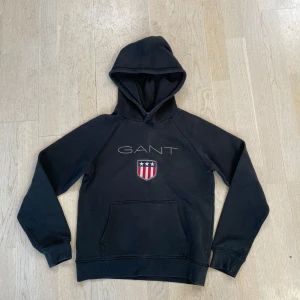 Svart hoodie från GANT med logga - Svart hoodie från GANT med broderad logga och sköld på bröstet. Klassisk känguruficka framtill och huva. Perfekt för en avslappnad stil.