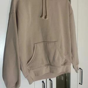 Beige hoodie med ficka - Säljer en enkel beige hoodie med huva. Tröjan har en stor magficka framtill och ribbade muddar vid ärmslut och nederkant. Perfekt för en avslappnad stil.