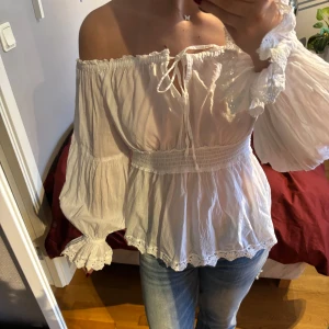 Vit offshoulder blus med spetsdetaljer - Säljer en vit offshoulder blus med långa vida ärmar och spetsdetaljer vid ärmslut och nederkant. Blusen har knytning framtill och smock i midjan för en feminin siluett. Perfekt till jeans eller kjol! Passar även S