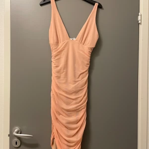 Oh Polly klänning - Säljer en persikofärgad tight klänning från Oh Polly med djup v-ringning. Klänningen har rynkade detaljer framtill och är tillverkad i ett stretchigt material som sitter snyggt på kroppen. Helt ny. 