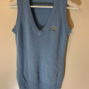 Ljusblå stickad väst från Gina Tricot - Ljusblå stickad väst från Gina Tricot med v-ringning och broderad gulddetalj på bröstet. Perfekt att styla över en skjorta eller t-shirt för en trendig look.