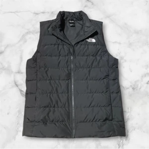 Svart dunväst från The North Face - Säljer en svart dunväst från The North Face med dragkedja framtill och ståkrage. Västen har en stilren design med logga på bröstet och ryggen. Perfekt för lager-på-lager och enkel att matcha med olika outfits. Nypris 1800kr.