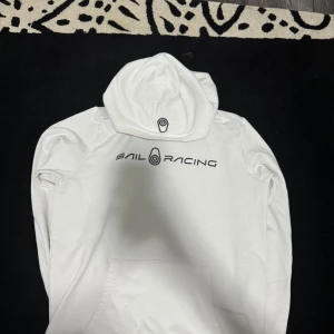 Vit hoodie från Sail Racing - Säljer en vit hoodie från Sail Racing med svart logga tryckt på bröstet och huvan. Tröjan har en klassisk känguruficka framtill och är tillverkad i mjukt material. Perfekt för dig som gillar stilrena och sportiga plagg.