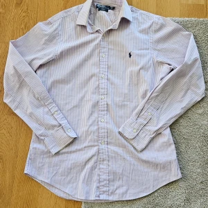 Ljuslila och vit randig skjorta från Ralph Lauren - Klassisk skjorta från Ralph Lauren i ljuslila och vitt med smala ränder. Skjortan har lång ärm, knappar framtill och broderad logga på bröstet. Perfekt till dig som gillar stilrena och tidlösa plagg.