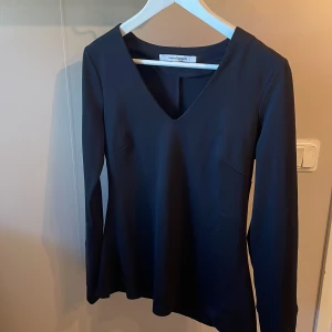Svart långärmad blus från KappAhl - En stilren svart långärmad blus från KappAhl med djup v-ringning. Blusen har en enkel, elegant design utan synliga detaljer eller mönster, vilket gör den lätt att styla till olika tillfällen.