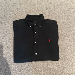 Svart skjorta från Ralph Lauren - Svart linne skjorta från Ralph Lauren med knappar framtill och röd broderad logga på bröstet. Skjortan har button-down krage och är perfekt för en stilren look. Använd typ 5 gånger. Nypris 1800 priset går att diskutera vid snabb affär 