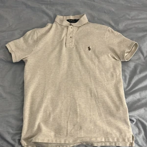 Ljusgrå pikétröja från Polo Ralph Lauren - Säljer en ljusgrå pikétröja från Polo Ralph Lauren med klassisk krage och knappar framtill. Tröjan har korta ärmar och den ikoniska broderade loggan på bröstet. Perfekt för en stilren och avslappnad look.