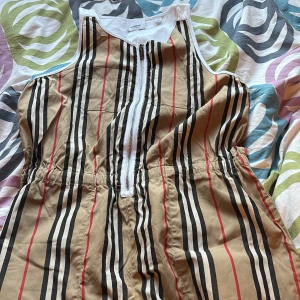 Randig ärmlös klänning från Burberry - Säljer en snygg ärmlös klänning från Burberry med klassiskt beige, svart och rött randigt mönster. Klänningen har dragkedja framtill och resår i midjan för en bekväm passform. Perfekt för dig som gillar stilrena och ikoniska plagg.