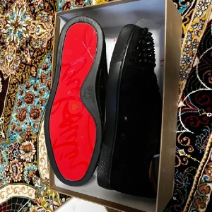 Louboutin skor - Säljer ett par svarta sneakers med coola svarta nitar framtill och klassisk röd sula. Skorna har snörning och är tillverkade i mocka och skinn. Perfekta för dig som vill sticka ut med en edgy stil.