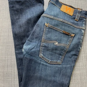 Blå jeans från Nudie Jeans - Säljer ett par klassiska blå jeans från Nudie Jeans med snygga slitningar och kontrastsömmar. Jeansen har fem fickor, normal passform och är tillverkade i ekologisk denim. Perfekta för dig som gillar en tidlös stil.