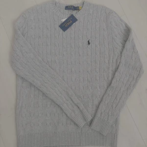 Ljusgrå stickad tröja från Polo Ralph Lauren - Säljer en ljusgrå stickad tröja från Polo Ralph Lauren med klassiskt kabelstickat mönster och broderad logga på bröstet. Tröjan har rund halsringning och långa ärmar. Tillverkad i 100% bomull. S men passar bättre m