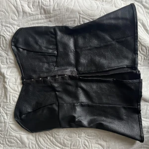 Svart korsett-topp i skinnimitation - Säljer en svart korsett-topp i skinnimitation. Toppen har knäppning framtill och en lätt glansig yta med struktur. Perfekt för dig som vill ha en edgy och trendig look.