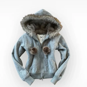 Grå hoodie från Abercrombie & Fitch - Säljer en grå hoodie från Abercrombie & Fitch med fuskpälsfodrad huva och pom-pom detaljer. Den har dragkedja framtill och ribbade muddar vid ärmslut och nederkant. Perfekt för kyliga dagar! Bud gjärna ett pris, köpte secondhand, aldrig använt själv men ser inga synliga defekter förutom en pytte liten ljus fläck på märket.Storlek L men passar mindre storlekar, den sitter inte som en L på mig som har oftast storlek S.  kedjan är lite trög men det är inga synliga defekt