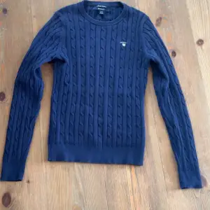 Säljer en stilren mörkblå kabelstickad tröja från GANT. Tröjan har en klassisk rund halsringning och ett diskret broderat märke på bröstet. Perfekt för kyligare dagar och ger en tidlös look. Det står storlek xs men passar dig som är 154-160