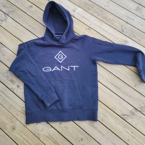 Mörkblå hoodie från GANT - Säljer en snygg mörkblå hoodie från GANT med deras ikoniska logga på bröstet. Tröjan har en klassisk känguruficka och en bekväm huva. Perfekt för en avslappnad stil.