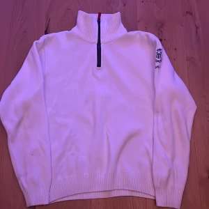 Vit tröja med dragkedja från Henri Lloyd - Säljer en stilren vit tröja från Henri Lloyd med halv dragkedja och broderad logga på ärmen. Tröjan har en hög krage och ribbade muddar. Perfekt för kyligare dagar.