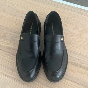 Svarta loafers från Copenhagen Shoes - Stilrena svarta loafers i skinn från Copenhagen Shoes. De har en klassisk design med en dekorativ detalj i guld på ovansidan. Perfekta för en elegant look.