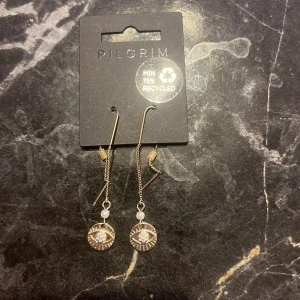 Guldörhängen med kedja och pärlor från Pilgrim - Eleganta guldörhängen från Pilgrim med en tunn kedja och små vita pärlor. De har en rund, glittrig hängande detalj som ger en stilren look. Perfekta för att lyfta vilken outfit som helst.