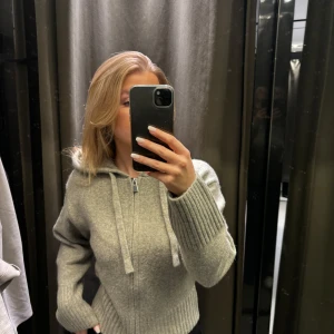 Grå stickad hoodie med dragkedja - Säljer en mysig grå stickad hoodie från Zara. Den har en dragkedja framtill och en justerbar huva med snören. Perfekt för kyliga dagar. Ribbstickade muddar vid ärmslut och nederkant ger en snygg look.