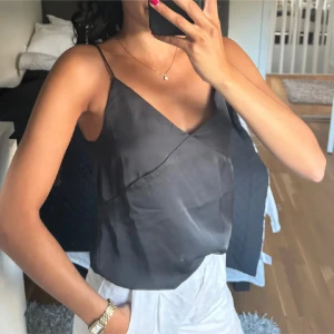 Svart satinlinne med smala axelband - Snyggt svart linne i satin med smala axelband och omlott-design. Perfekt för en stilren look. Linnet har en elegant knytning i ryggen som ger en extra detalj.