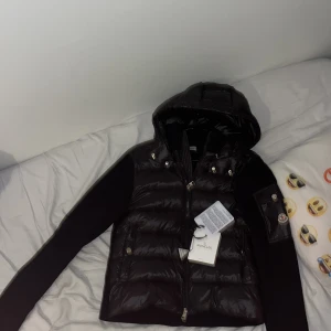 Svart dunjacka från Moncler - Snygg svart dunjacka från Moncler med huva och dragkedja. Jackan har ett stilrent märke på ärmen och är perfekt för kyligare dagar. Den är långärmad och har en glansig finish som ger en exklusiv look.