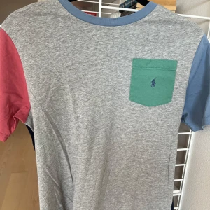 Flerfärgad t-shirt från Ralph Lauren - Säljer en cool flerfärgad t-shirt från Ralph Lauren. Den har en grå framsida med en grön bröstficka och blå logga, samt blå och röda ärmar. Baksidan är mörkblå. Perfekt för en avslappnad stil!