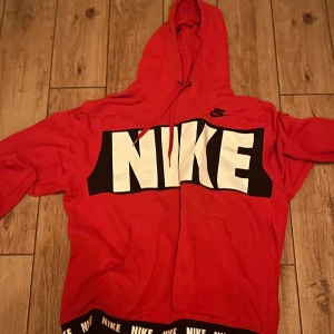 Röd hoodie från Nike - Säljer en röd hoodie från Nike med stor logga framtill. Tröjan har en justerbar huva och långa ärmar. Perfekt för en sportig look eller avslappnade dagar.